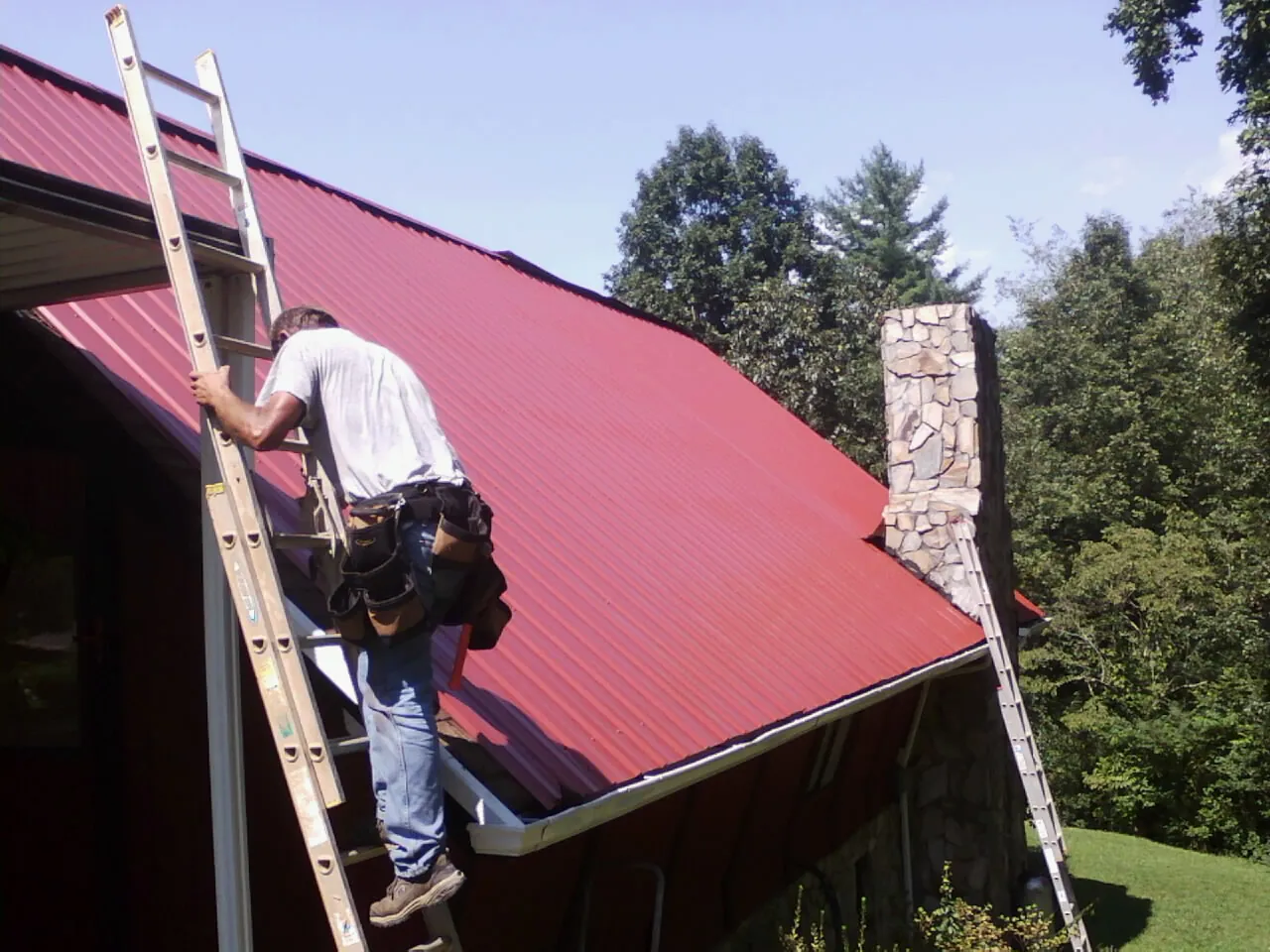 Roofer on a ladder beside a red metal roof and stone chimney — Soffit & Fascia Repair by Roofing Brothers in Bon Air